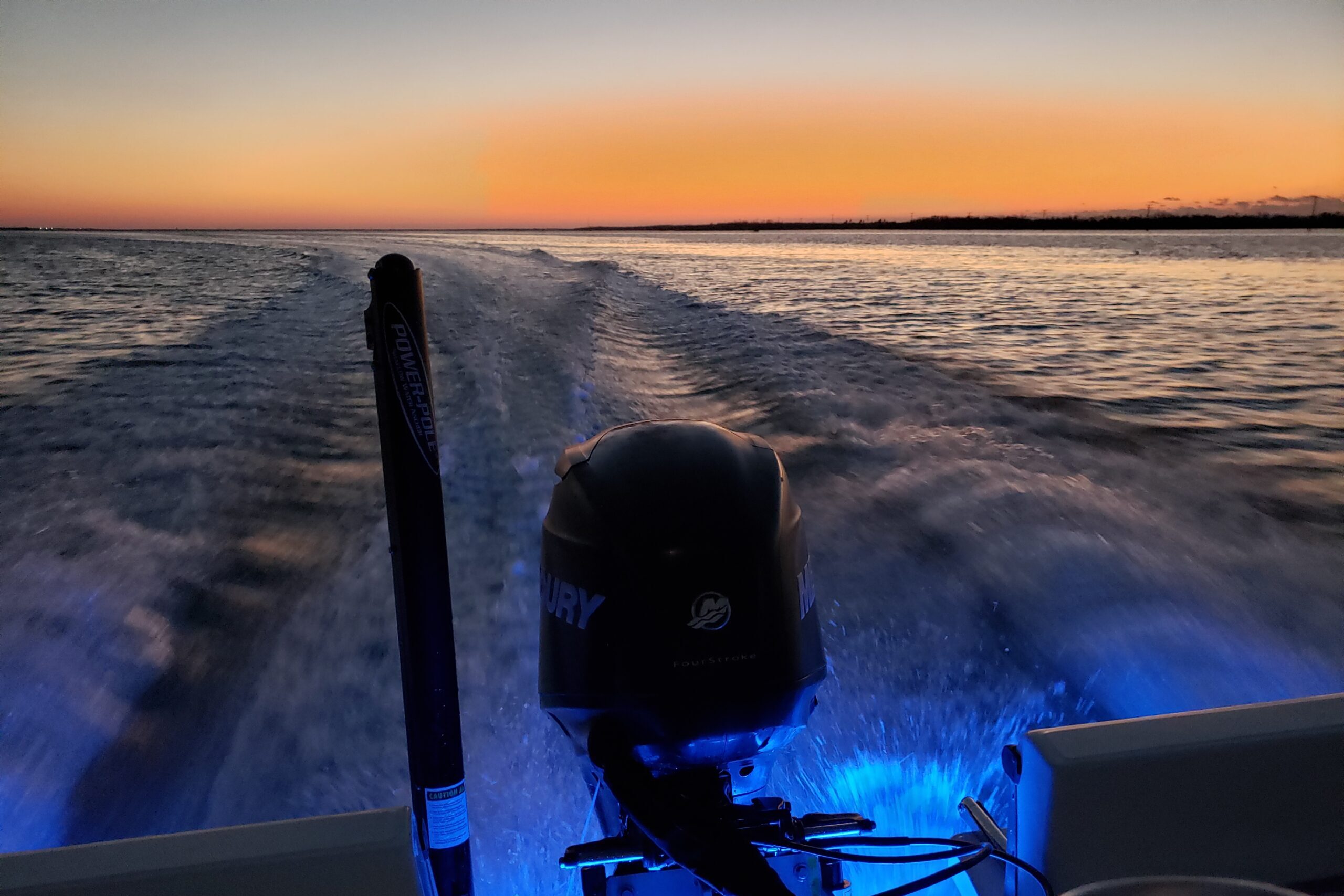 Mercury engine on a boat glowing with blue underwater lights during a nighttime cruise through the Gulf of Mexico at sunset.