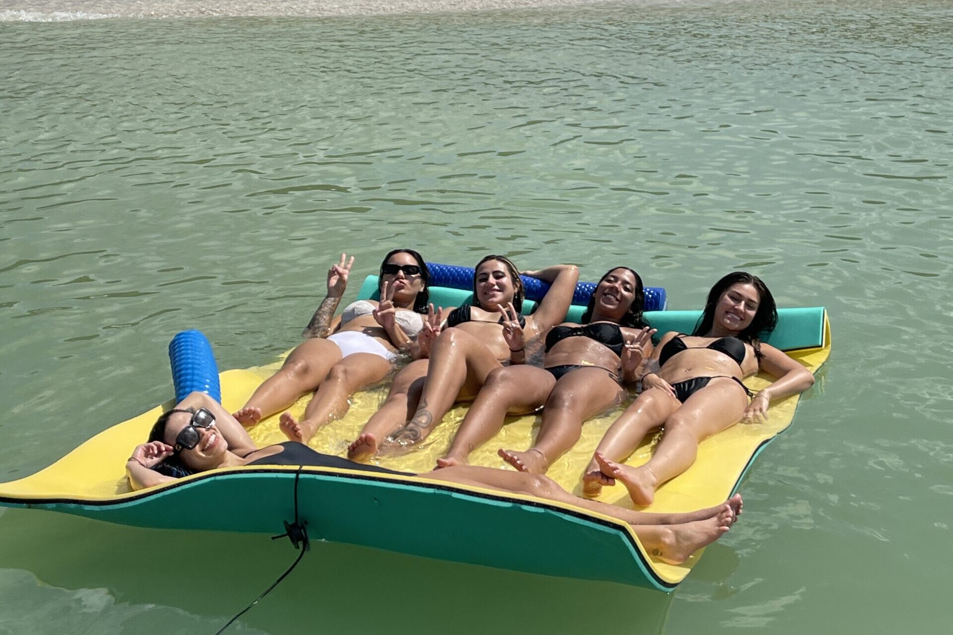 Five women in swimsuits smiling and throwing peace signs while lounging together on a floating mat near a beach in Naples, Florida.