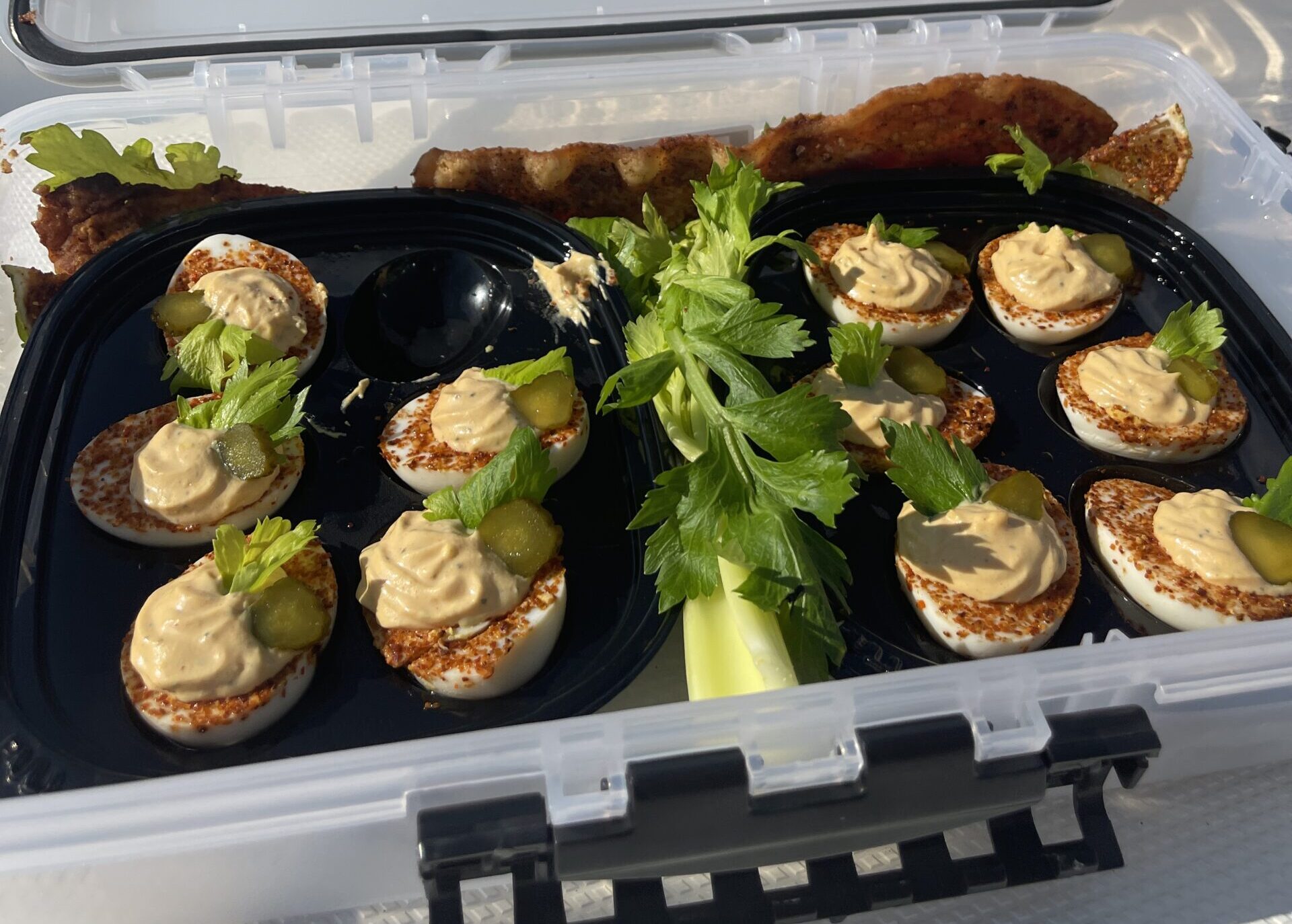 Waterproof tackle box filled with Bloody Mary-style deviled eggs topped with pickles and celery, served with crispy bacon and celery stalks on a boat at the shoreline