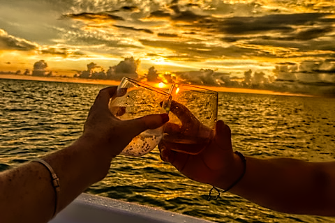 Two hands clinking cocktail glasses at sunset on a boat with golden skies and calm ocean water in the background.